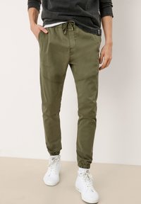 Man in olijfgroene joggers met een trekkoord in de taille en elastische boorden, gecombineerd met witte hoge sneakers en een donkere lange mouw shirt.