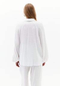 Weißes, strukturiertes, übergroßes Hemd mit einem kragenlosen Ausschnitt und langen, voluminösen Ärmeln, von hinten betrachtet. Glatter Stoff, lässige Passform.