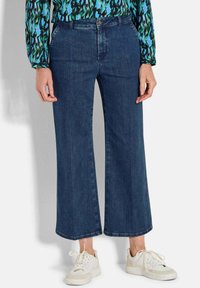 Weite, dunkelblaue Jeans mit hohem Bund und rohem Saum. Kombiniert mit weißen Sneakers. Die Textur wirkt robust mit einem glatten Finish.