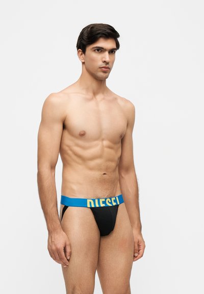 Jeune homme aux cheveux courts et foncés portant un jockstrap noir avec une ceinture bleue avec un texte jaune "DIESEL".