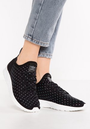 Person trägt schwarze Adidas-Sneaker mit irisierenden Punkten und weißen Sohlen, kombiniert mit blau umgeschlagenen Jeans.