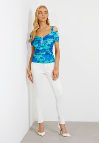 Blå blomsteret off-the-shoulder top med rynket design, parret med hvide tætsiddende jeans og hvide spidse hæle på en neutral baggrund.
