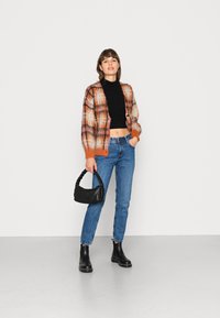 Cardigã xadrez multicolorido com textura suave, em tons de laranja e castanho, usado sobre um suéter preto com gola alta cropped. Combinado com jeans azuis e uma bolsa preta.