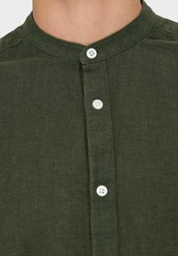 Chemise en lin vert foncé avec un col montant, fermeture boutonnée avec des boutons blancs, et un tissu doux et texturé.