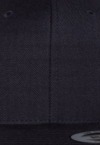 Gorra de béisbol de un oscuro color navy con una tela texturada, presenta un visera plana y un parche circular con el logo en la parte frontal, con detalles de costura en contraste.