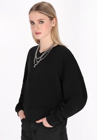 Fekete, oversized kapucnis pulóver V-nyakkivágással, hosszú ujjakkal és bordás aljjal. Rétegezett ezüstlánc nyakláncokkal viselve. Sima textúrájú.
