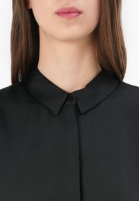 Zwarte blouse met kraag, gemaakt van een gladde stof en met een knoopsluiting. Dichtbijzicht legt de nadruk op de details van de halslijn en kraag.