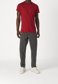 Hombre con una camiseta roja ajustada de manga corta, pantalones vaqueros holgados de color gris oscuro y zapatillas blancas, de pie frente a un fondo blanco liso.