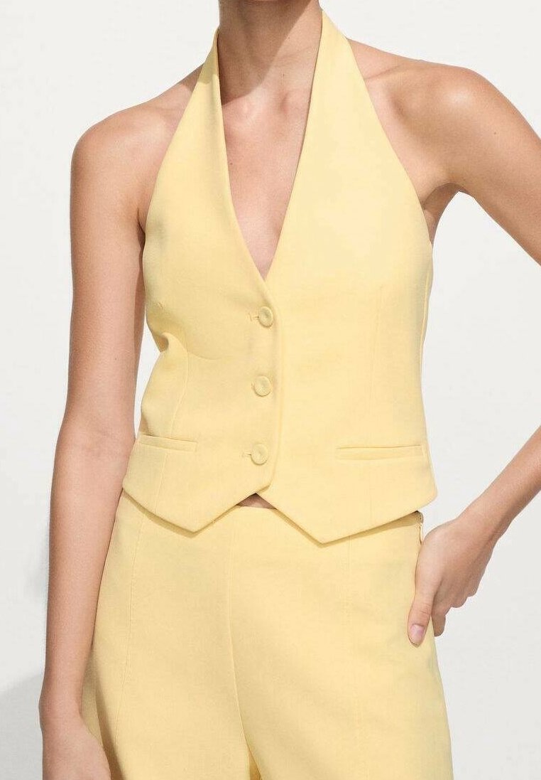 Femme portant un gilet halter jaune sans manches avec des boutons et un pantalon taille haute assorti, posant avec une main sur la hanche.