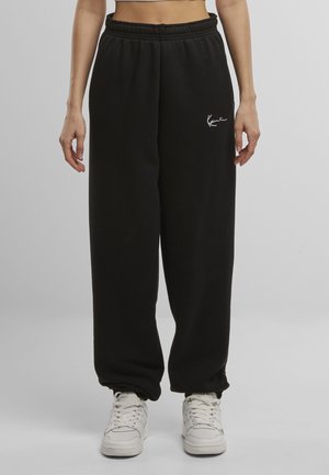 Fekete pamut melegítőnadrág, rugalmas derékrésszel, bő szabással és egy kis fehér logóval a bal comboldalon. Fehér tornacipővel viselve.