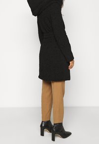 Svart strukturerad kappa med huva, bälte i midjan och långa ärmar. Kombineras med tan-färgade smalna byxor och svarta klackstövletter.