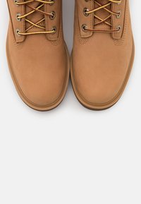 Bottes en suède beige avec bout rond, œillets en métal, lacets contrastants jaunes et rouges, et semelle en caoutchouc. Texture lisse et détails de couture minimalistes.