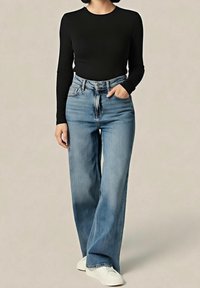 Femme portant une chemise noire à manches longues, un jean bleu taille haute à jambes larges, et des chaussures blanches, debout avec une main dans la poche.