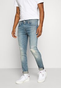 Ljusblå denimjeans med slim fit, med slitna lappar och uppvikta ärmslut, parat med vita sneakers och en enkel vit t-shirt.
