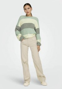 Egy nő áll, laza csíkos pulóvert visel mentazöld, szürke és krém színekben, bézs széles lábú nadrággal és krém színű sportcipővel.