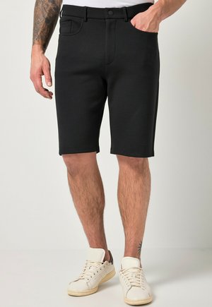 Mand iført sorte shorts i mid-længde og hvide sneakers, stående med den ene hånd i lommen mod en lys baggrund.