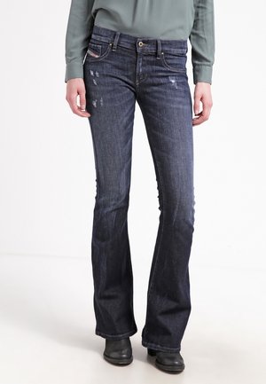 Nainen pukeutuneena tummansinisiin liehuviin farkkuihin, joissa on hieman kulutuksen jälkiä, vihreään pitkähihaiseen paitaan ja mustiin nilkkureihin, seisoo valkoisella taustalla.