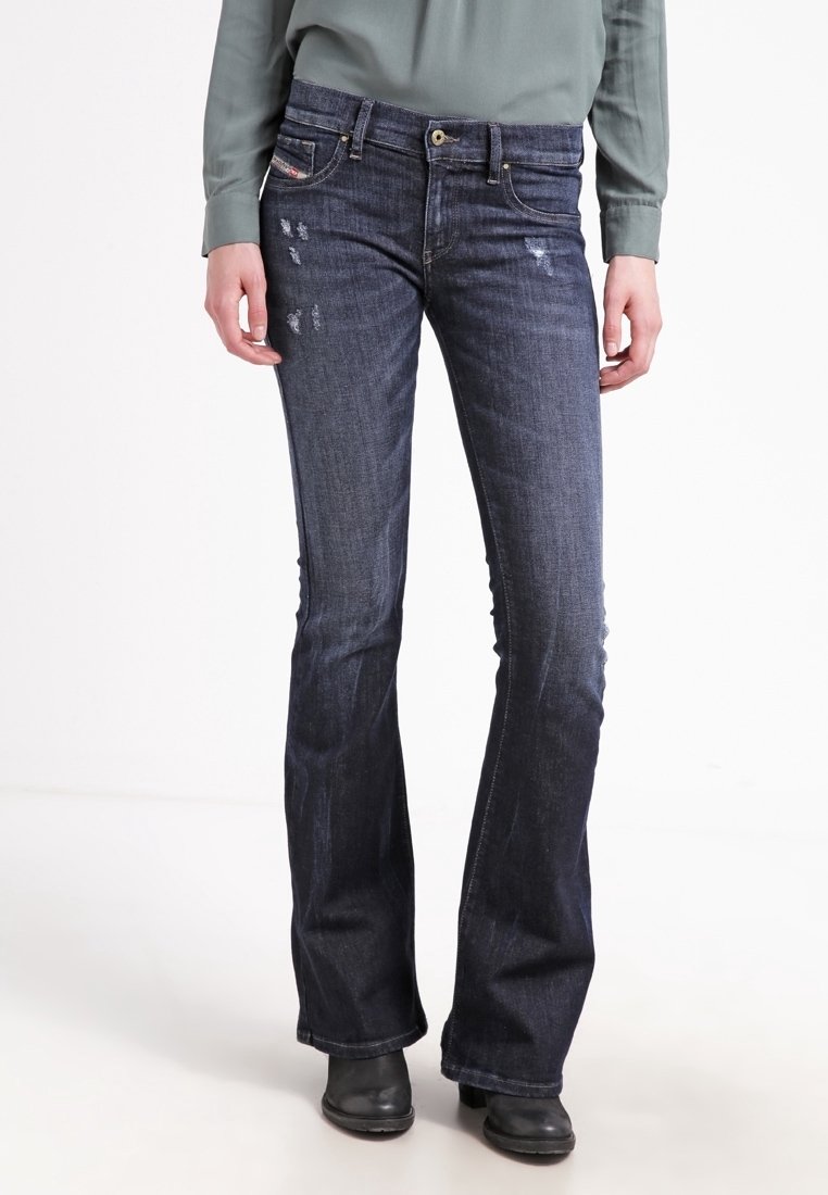 Femme portant un jean évasé bleu foncé légèrement usé, un chemisier vert à manches longues et des bottines noires, debout sur un fond blanc.