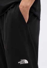 Persona che indossa pantaloni neri The North Face con la mano nella tasca laterale, logo bianco visibile sulla coscia, sfondo uniforme.