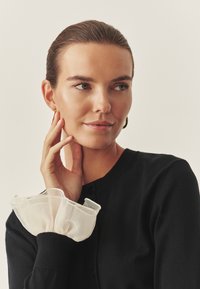 Crni kardigan s okruglim izrezom i dugmadi, s prozirnim bijelim čipkastim manžetama. Glatka tekstura tkanine i prilagođeni dizajn.