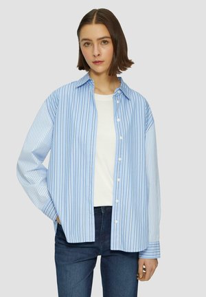 Chemise à rayures bleues et blanches, boutonnée, en tissu léger, dotée d'un col, de manches longues et d'une coupe décontractée. Portée par-dessus un t-shirt blanc.