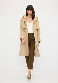 Beige trenchcoat med huva, två framfickor och tryckknappar. Matchad med olivgröna byxor och spetsiga gröna klackar.