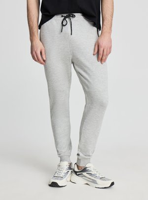 Mężczyzna w jasnoszarych spodniach dresowych z czarnym sznurkiem oraz białych butach do biegania, stojący na tle jednolitego jasnego tła.