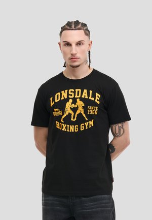 Giovane con i capelli intrecciati che indossa una t-shirt nera della Lonsdale Boxing Gym con stampa gialla e jeans scuri, in piedi davanti a uno sfondo grigio chiaro.