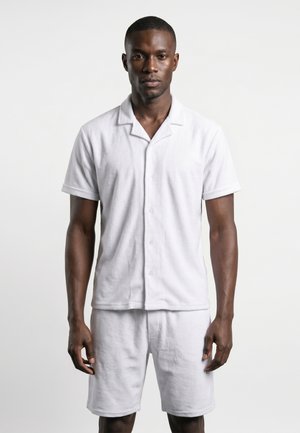 Homme debout, faisant face à l'avant, portant une chemise blanche à manches courtes boutonnée et un short blanc assorti sur un fond uni.