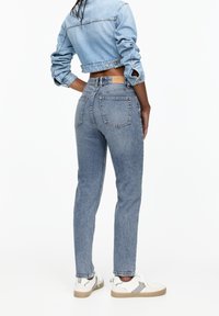 Femme portant une veste en jean courte bleu clair, un jean bleu ajusté à taille haute et des baskets blanches, debout devant un fond blanc.