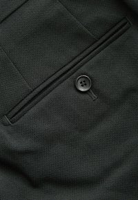 Črn teksturiran material z gladko površino, ki ima žep na zadrgo in okrasni črn gumb z obročkom in štirimi luknjami.