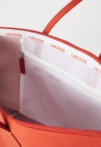 Röd tote-väska med en texturerad utsida, inuti vit mesh och en ficka med dragkedja med en röd etikett. Lacoste-logotyp längs kanten.