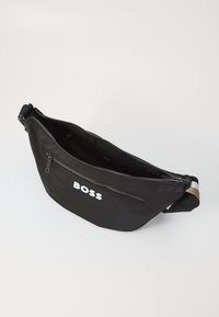 Sac banane en nylon noir avec fermeture éclair, doté d'une poche avant et d'un logo blanc. Il présente une finition texturée et une bandoulière réglable.