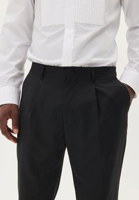 Melni svārki ar pieguļošu piegriezumu, gludu tekstūru un dubultām krokām priekšpusē. Saskaņoti ar baltu formālo krekli, kura plecu daļa ir krokota.