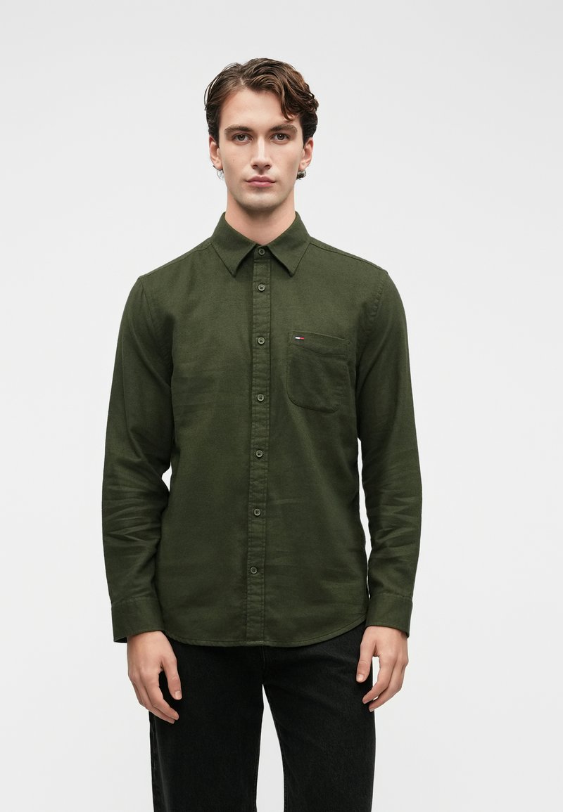 Σκοτεινά πράσινο πουκάμισο με κουμπιά, φτιαγμένο από βαμβάκι, διαθέτοντας τσέπη στο στήθος και ένα μικρό λογότυπο. Μακριά μανίκια με κουμπωτές μανσέτες.