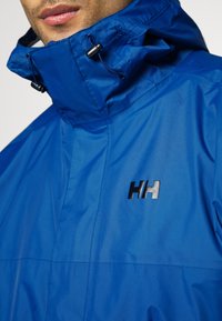 Chaqueta impermeable azul con cuello alto, ajustes de cordón y acento de logo negro. Tejido ligero con una textura suave.