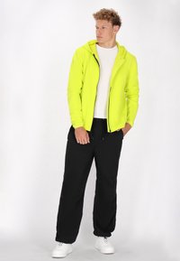 Neon gele ritshoodie met een aansluitend ontwerp, gecombineerd met losse zwarte broeken. Witte sneakers maken de outfit af. Glad, lichtgewicht materiaal.