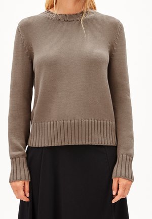 Femme portant un pull en maille taupe avec poignets et ourlet côtelés, associé à une jupe noire, debout devant un fond blanc uni.
