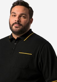 Svart polo tröja med gul krage och fickkant, som har en knäppning framtill och en mjuk bomullskvalitet, skräddarsydd för en avslappnad passform.