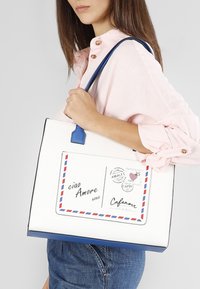 Vit tote bag med blå detaljer har postkortgrafik, röd och blå kuvertkant och svart text; tillverkad av syntetiskt material.