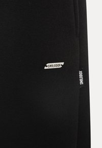 Musta kangas, jossa on pieni suorakulmainen metallilätkä ja kangasmerkki, joissa molemmissa on brändin nimi "Smilodox".