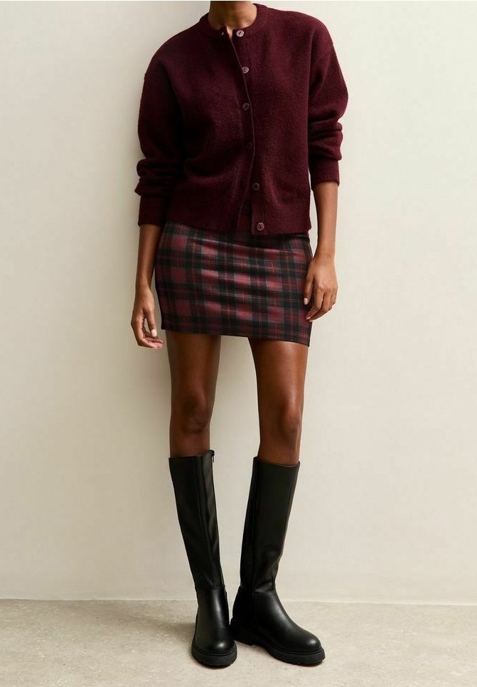 Femme portant un cardigan bordeaux boutonné, une mini-jupe à carreaux rouges et noirs, et des bottes noires jusqu'aux genoux, debout contre un mur beige.