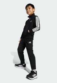 Černý sportovní teplákový komplet s bílými pruhy, zipovou bundou a odpovídajícími kalhotami. V páru s černými teniskami s třemi bílými pruhy.