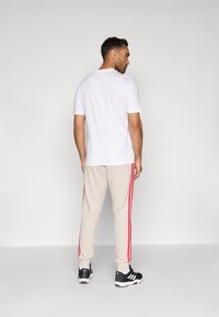 Balta trumpa rankovėmis marškinėliai kartu su smėlio spalvos džinsais, turinčiais raudonas šonines juostas ir juodus sportinius batus. Paprastas fonas.