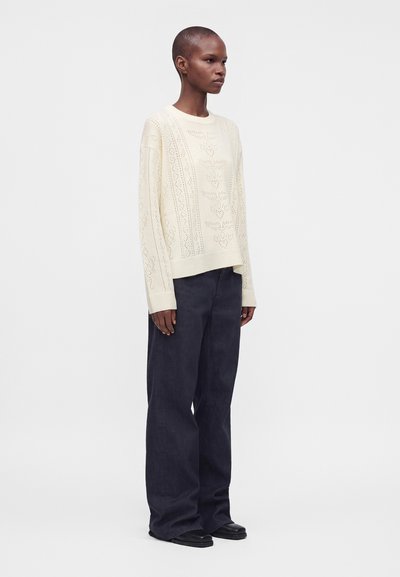 Personne aux cheveux courts portant un pull en maille crème à motifs et un pantalon large sombre, debout dans un espace neutre.