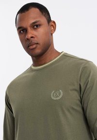 Uomo con capelli corti che indossa una camicia a maniche lunghe verde oliva con un piccolo logo EX ricamato sul petto, che guarda avanti.