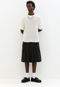 Maglione incurvato a righe bianche, pantaloni corti neri con texture, calze bianche e scarpe stringate nere. Accento con una semplice collana d'oro.