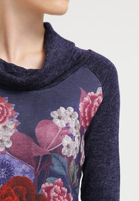 Detailní záběr osoby v tmavě modrém svetru s květinovým vzorem a měkkým, volně splývajícím límcem.