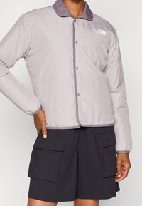 Ljusgrå puffjacka med lila krage, tryckknappar och sidofickor, kombinerad med mörka cargoshorts. Slät textur och avslappnad design.