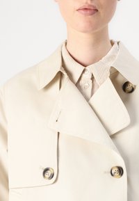 Trench coat beige con colletto a punte, design doppiopetto e grandi bottoni circolari. Camicia chiara a trama sotto.